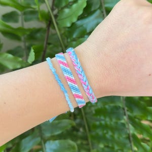 May include: Four handmade friendship bracelets in shades of blue, pink, and white are displayed on a wrist. The bracelets feature various designs, including a braided style and a simple twisted cord. The background is a lush green foliage.