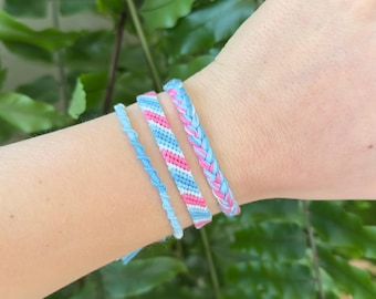 Puede incluir: Cuatro pulseras de amistad hechas a mano en tonos de azul, rosa y blanco se muestran en una muñeca. Las pulseras presentan varios diseños, incluido un estilo trenzado y un simple cordón retorcido. El fondo es un exuberante follaje verde.