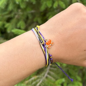 May include: A bracelet with multiple strands in black, yellow, white, and purple. It has an orange carriage charm with gold details. The bracelet is tied on the wrist.