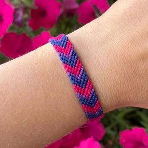 Peut inclure: Un bracelet d'amitié tissé présentant un motif chevron répétitif dans des tons alternés de bleu foncé, de violet et de rose vif. Le bracelet est fabriqué à partir de fil à broder fin et étroitement noué, créant une surface texturée.
