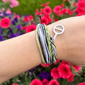 Peut inclure: Un bracelet composé de plusieurs brins de fils verts, gris, noirs et blancs. Un charm argenté avec un cercle barré est attaché. Le bracelet est présenté sur un fond de fleurs colorées.