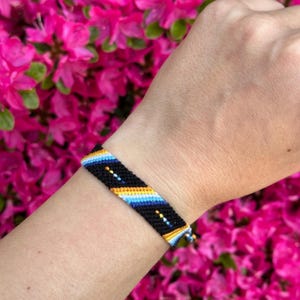 May include: A black woven friendship bracelet with yellow, blue, and white stripes. The bracelet is worn on a wrist, set against a background of pink flowers.