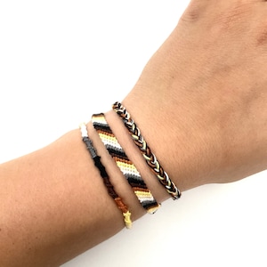 Peut inclure: Quatre bracelets d'amitié de couleur marron, noir, blanc et jaune. Les bracelets sont tissés selon un motif simple et sont attachés au poignet d'une personne.