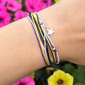Peut inclure: Un bracelet à plusieurs brins avec des cordons blancs, jaunes, noirs et violets. Il comporte un petit charme en forme d'étoile argentée et des perles argentées. Le bracelet est noué avec une ficelle violette.