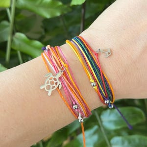 Peut inclure: Gros plan sur deux bracelets d'amitié colorés. L'un des bracelets présente un charm en forme de tortue argentée, l'autre un charm en forme d'ancre argentée. Les bracelets sont faits de plusieurs brins de fil de différentes couleurs, dont le rose, l'orange, le jaune, le vert et le bleu.