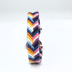 May include: A woven friendship bracelet with a chevron pattern. The bracelet displays a range of colours, including orange, white, blue, pink, black, and purple. It is made of thread and has a knotted fastening. The bracelet is approximately 1.5 cm wide.