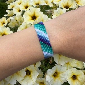 May include: A woven friendship bracelet with a diagonal pattern in shades of green, purple, and white.