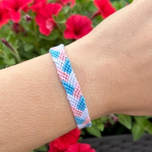 Pode incluir: Uma pulseira de amizade tecida com um padrão de chevron. A pulseira apresenta um esquema de cores azul claro, rosa e branco. A pulseira é usada em um pulso, com flores vermelhas no fundo.