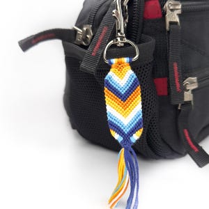 May include: A woven chevron-patterned keychain featuring orange, white, light blue, and dark blue threads, attached to a silver-toned metal lobster clasp. The keychain ends in a multi-colored tassel made of loose threads in matching colors.