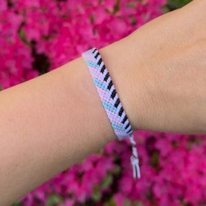 May include: Woven friendship bracelet in pastel colors. The bracelet features a chevron pattern with light blue, pink, and black threads. The bracelet is on a wrist with a blurred background of pink flowers.