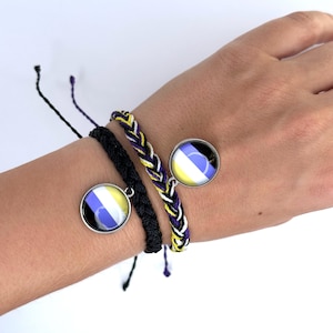 May include: Two bracelets with black and white braided cords and a charm featuring a yellow, white, and purple circle design. The bracelets are worn on a person's wrist.