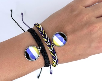 May include: Two bracelets with black and white braided cords and a charm featuring a yellow, white, and purple circle design. The bracelets are worn on a person's wrist.