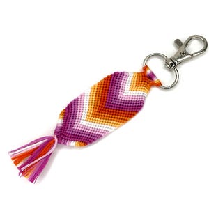 Könnte beinhalten: Ein handgefertigter Schlüsselanhänger mit einem gewebten Design in den Farben der lesbischen Pride-Flagge: Orange, Rosa und Weiß. Der Schlüsselanhänger hat einen silberfarbenen Verschluss und eine Quaste aus passenden Fäden.