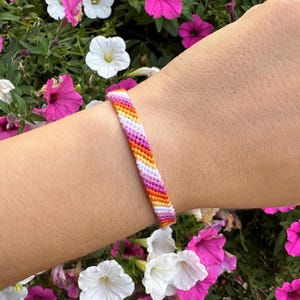 May include: A colourful woven bracelet with a chevron design, showing stripes of orange, yellow, white, pink, and purple. The bracelet is set against a background of pink and white petunia flowers.