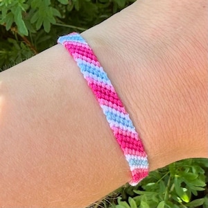 Puede incluir: Una pulsera de amistad hecha a mano con un diseño de rayas. La pulsera presenta rayas alternas rosas, celeste y blancas. La pulsera se lleva en una muñeca, con follaje verde en el fondo.