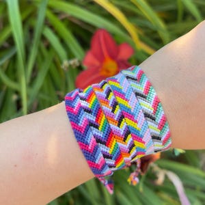 May include: A wide woven friendship bracelet featuring a repeating chevron pattern in vibrant rainbow colours including purple, blue, green, yellow, orange, and red, alongside a secondary section with pastel pink, blue, and white tones.