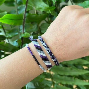 Op de afbeelding: Vier handgemaakte vriendschapsarmbanden in tinten paars, zwart, grijs en wit worden op een pols getoond. De armbanden hebben verschillende geweven patronen, waaronder strepen en vlechten. De achtergrond is een weelderige groene plant.
