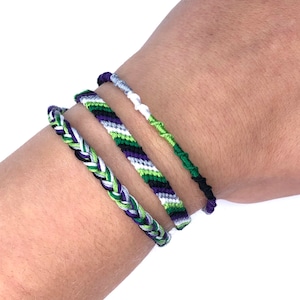 May include: Three friendship bracelets in green, white, black, and purple colors. The bracelets are woven together in a braided style.