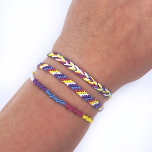 May include: Three friendship bracelets in different colors and patterns. The bracelets are made of woven string and feature a variety of colors, including yellow, white, purple, blue, and red.