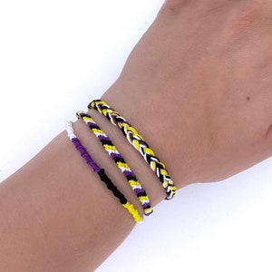 May include: Four friendship bracelets in yellow, purple, black, and white colours. The bracelets are braided and are worn on a person's wrist.