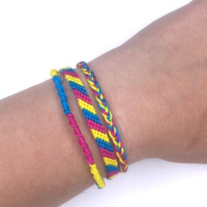 May include: Four friendship bracelets in yellow, blue, and pink colors. The bracelets are woven with a variety of patterns, including chevron, stripes, and a simple braid.
