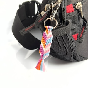 Op de afbeelding: Een zwarte heuptas met een ritssluiting en een kleurrijke geweven sleutelhanger eraan. De sleutelhanger is roze, oranje, blauw en wit.