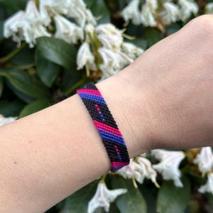 May include: A woven friendship bracelet in the colours of the bisexual pride flag: pink, purple, and blue. The bracelet is black with diagonal stripes of pink, purple, and blue. The bracelet is worn on a wrist.