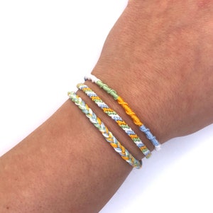 May include: Three braided friendship bracelets in yellow, green, and blue colors. The bracelets are worn on a person's wrist.