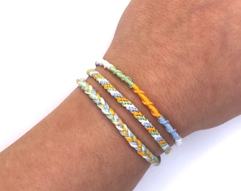 May include: Three braided friendship bracelets in yellow, green, and blue colors. The bracelets are worn on a person's wrist.