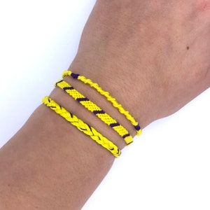 May include: Three yellow friendship bracelets with purple stripes on a person's wrist. The bracelets are made of woven string.