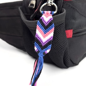 Può includere: Un portachiavi a braccialetto dell'amicizia intrecciato con un motivo a chevron nei toni del viola, rosa, blu, bianco e nero. Il ciondolo in tessuto è fissato a un moschettone in metallo argentato e pende dal lato di un marsupio in nylon nero.