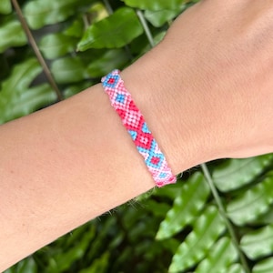 Op de afbeelding: Een handgemaakte vriendschapsarmband in roze, rood en blauw. De geweven armband heeft een geometrisch patroon. De armband wordt getoond tegen een achtergrond van groen gebladerte.