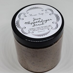 Puede incluir: Un tarro de Java Whipped Sugar Scrub de The Mosaic Soap Co. La etiqueta incluye el nombre del producto, los ingredientes y el peso neto de 142 gramos. El exfoliante es de color marrón claro y está contenido en un tarro transparente con tapa negra.