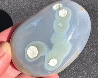 May include: A smooth, gray and white agate stone with a unique pattern of white, flower-like inclusions. The stone is polished and has a rounded shape.
