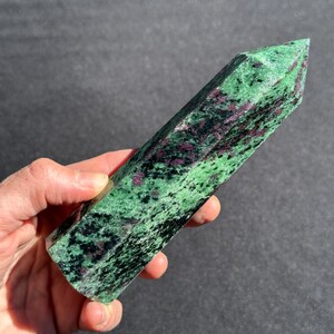 May include: A polished ruby zoisite crystal point. The stone features a vibrant green base color with black and deep red inclusions. The crystal is held in a hand, showcasing its natural beauty and unique patterns. This gemstone is a decorative item.