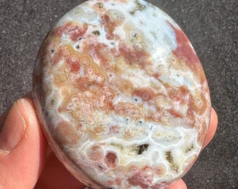 Può includere: Una pietra levigata di forma ovale con un motivo unico di cerchi concentrici e vortici. La pietra presenta una miscela di colori, tra cui bianco, rosso e marrone, creando un effetto marmorizzato. La pietra è liscia e sembra essere una pietra preziosa naturale.
