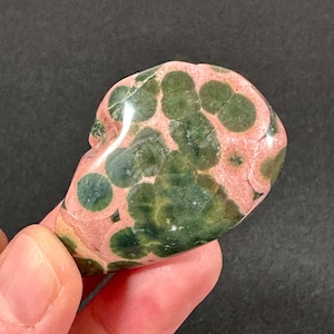 May include: A polished unakite stone with a mottled pattern of green and pink. The stone has a smooth, rounded shape and is held in a hand. The unakite is a metamorphic rock, often used in jewelry and decorative items.