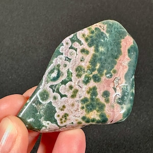 May include: A polished gemstone with a unique pattern of green, pink, and white. The stone has a smooth, rounded shape and a glossy finish. The colors are distributed in a mottled pattern, creating a visually interesting and natural appearance.