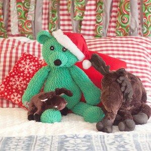 May include: Three plush stuffed animals: a green teddy bear wearing a red and white Santa hat, a brown moose, and a smaller brown reindeer. The toys are sitting on a white and red gingham blanket.