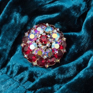 Puede incluir: Una tela de terciopelo azul turquesa con un broche grande, redondo, de diamantes de imitaci&oacute;n rojos y transparentes.