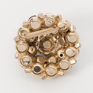 Puede incluir: Un broche dorado con un grupo de piedras de imitaci&oacute;n rojas. El broche tiene un cierre de pin y est&aacute; dise&ntilde;ado en forma de flor.