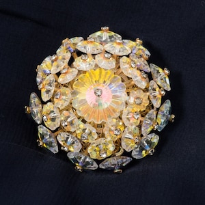May include: A large, round brooch made of gold-toned metal and clear iridescent glass. The brooch is shaped like a cluster of flowers with a central flower that is larger than the others.