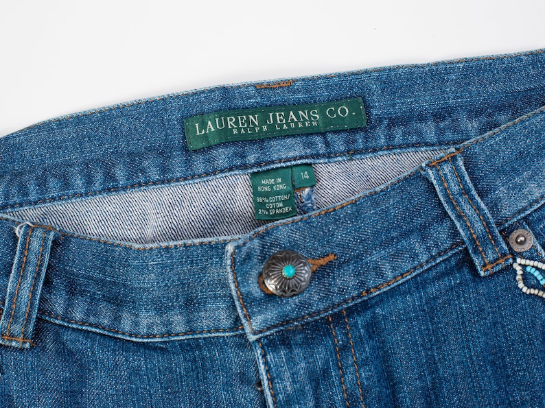 May include: Blue denim jeans with a green label that reads "Lauren Jeans Co. Ralph Lauren" and a size tag that reads "14". The jeans have a turquoise button and a turquoise and white beaded embellishment.