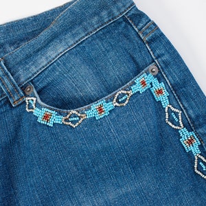 May include: Blue denim jeans with a beaded design around the pocket and side seam. The beads are turquoise, white, and red.