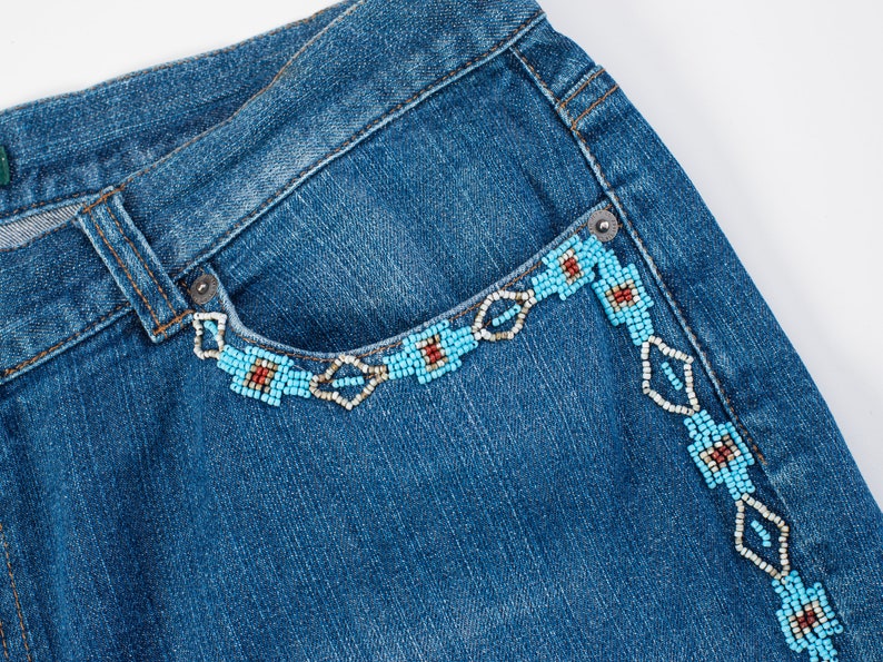 May include: Blue denim jeans with a beaded design around the pocket and side seam. The beads are turquoise, white, and red.