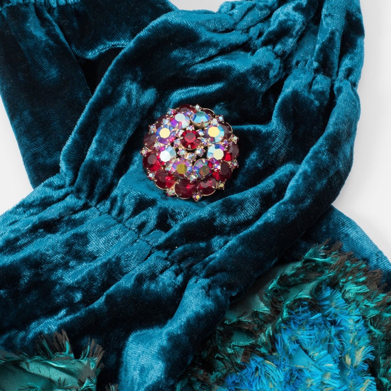 Puede incluir: Una tela de terciopelo azul turquesa con un broche grande, redondo, de diamantes de imitaci&oacute;n rojos y transparentes.
