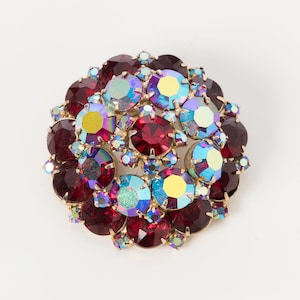 Op de afbeelding: Een cluster van rode, blauwe en iriserende strass stenen gezet in goudkleurig metaal. De strass stenen zijn in een cirkelvormig patroon gerangschikt en creëren een sprankelend effect.