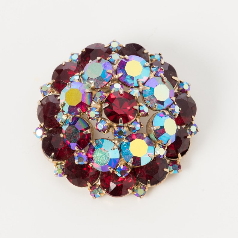 Puede incluir: Un grupo de piedras de cristal rojo, azul e iridiscente engastadas en metal dorado. Las piedras de cristal est&aacute;n dispuestas en un patr&oacute;n circular y crean un efecto brillante.