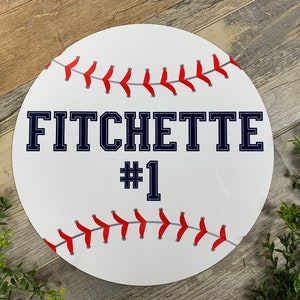 May include: White baseball-themed round sign with red stitching and the text "FITCHETTE #1" in navy blue.
