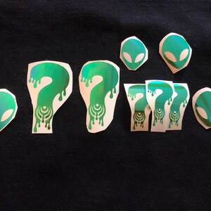 May include: A set of nine green and white iron-on patches featuring alien heads and question marks. The patches are perfect for adding a unique touch to clothing, bags, or other accessories.